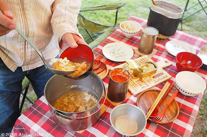 コーンスープをよそう人、缶詰料理、飯盒、食器、キャンプテーブル、チェア